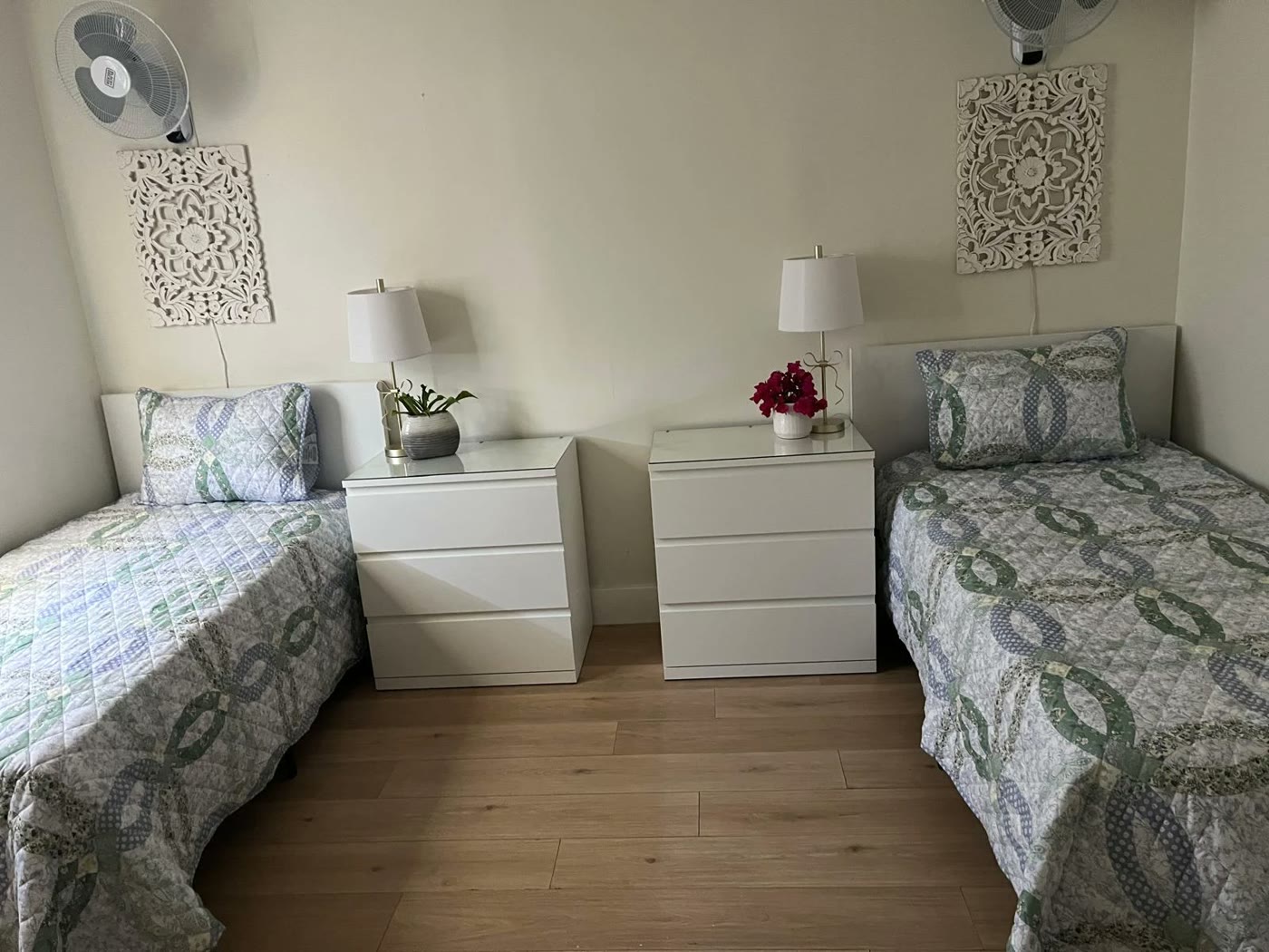 A shared bedroom: two made beds with quilted covers, white chests of drawers, lamps and fresh flowers.