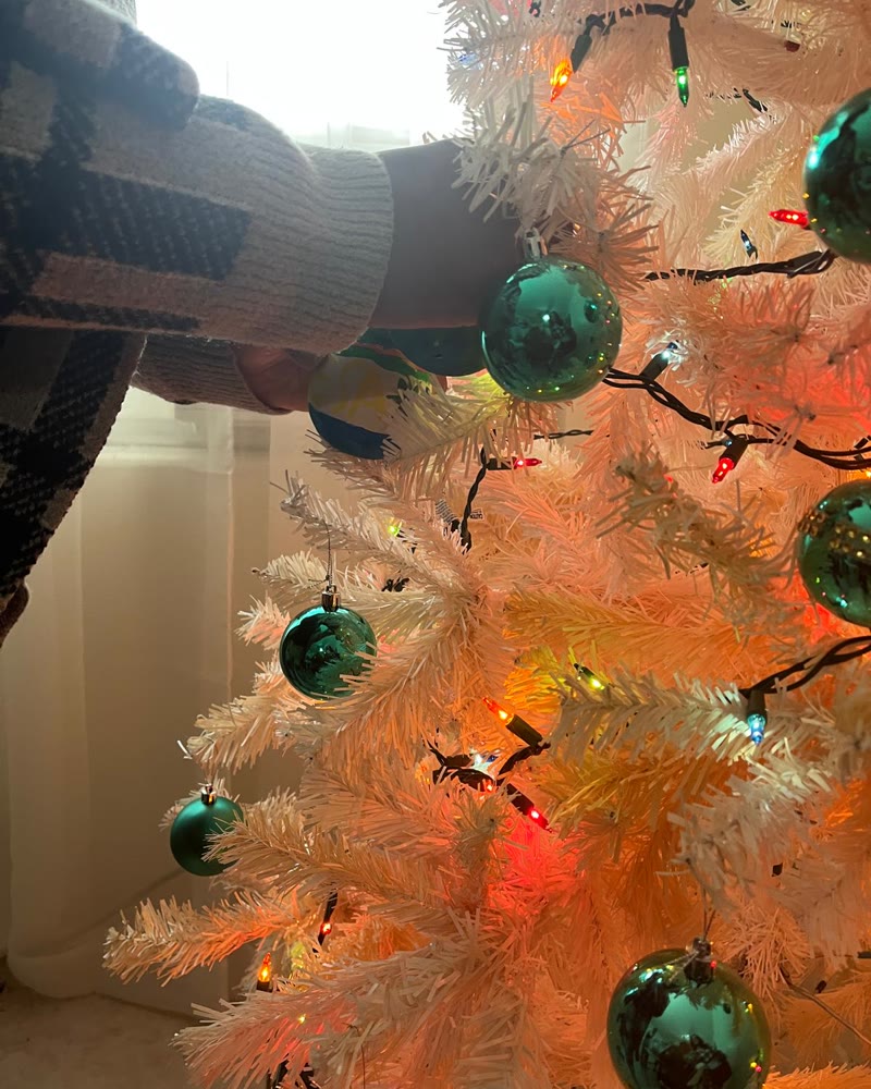 A hand hanging a green ornament on a white tree strung with colored lights.